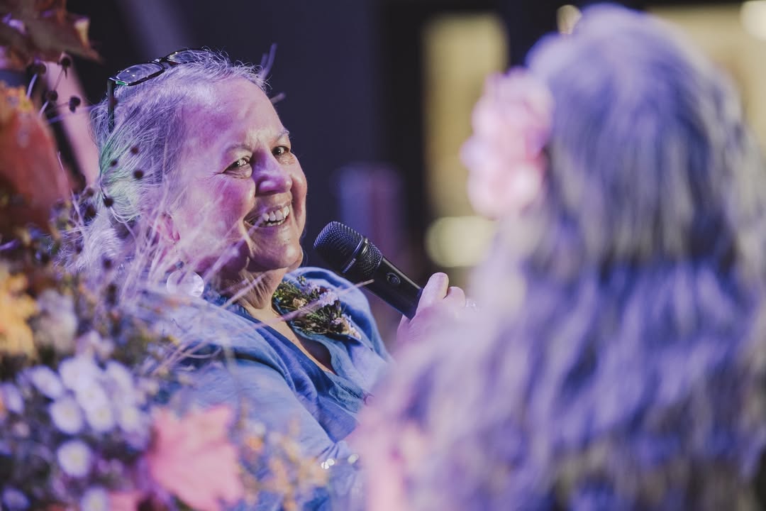 Robin Wall Kimmerer smiling and speaking into a microphone, framed by flowers. Robin Wall Kimmerer smiling and speaking into a microphone, framed by flowers.